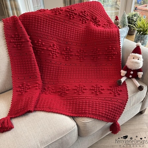 May include: A vibrant red crocheted blanket with a snowflake pattern, draped over a light gray sofa. The blanket features textured details and is adorned with tassels. A Santa Claus doll sits on the sofa. The blanket is a cozy home decor item.