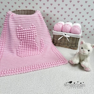 Puede incluir: Una manta de bebé de crochet rosa con un diseño de corazón texturizado. La manta está sobre una superficie blanca con una cesta de mimbre llena de hilo rosa y un juguete de peluche de gato blanco.