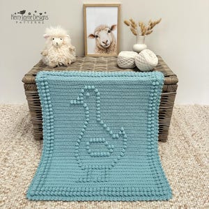 May include: A light blue crochet blanket featuring a textured bobble stitch duck motif in the centre and a matching bobble stitch border. The blanket is rectangular with a uniform stitch pattern throughout, showcasing a clean and simple design.