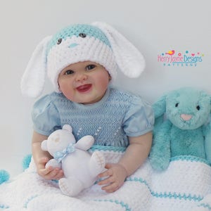 May include: A baby wearing a blue and white crocheted bunny hat and a blue smocked dress is sitting on a white and blue crocheted blanket with a blue bunny and a white teddy bear. The blanket has blue pom-poms on the edges.