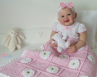 May include: A pink crocheted baby blanket with white flowers. The blanket has a white crocheted border. A baby doll is sitting on the blanket holding a white teddy bear.