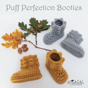 May include: Two pairs of crocheted baby booties, one pair in mustard yellow and one pair in gray. The booties have a puffy texture and a ruffled cuff. The booties are photographed on a white background with a branch of oak leaves and acorns.