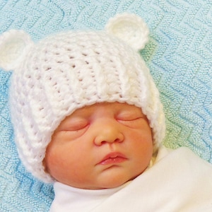 Pode incluir: Um chapéu de bebé branco de crochê com orelhas de urso. O chapéu está na cabeça de um bebé que está a dormir. O fundo é um cobertor azul claro.
