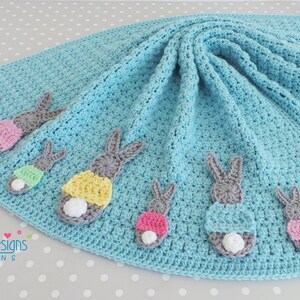 May include: A light blue crocheted baby blanket with gray bunny appliqu&eacute;s, each with a colorful skirt and a white fluffy tail. The blanket has blue pom-poms at the corners. The blanket is a handmade item.