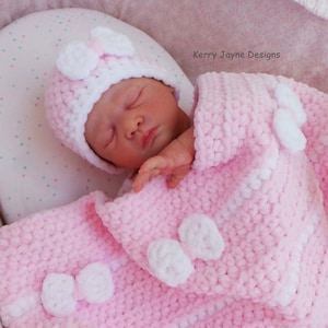 K&ouml;nnte beinhalten: Eine rosa geh&auml;kelte Babydecke mit wei&szlig;en geh&auml;kelten Blumen und Schleifen. Ein Baby schl&auml;ft auf der Decke und tr&auml;gt eine rosa und wei&szlig;e geh&auml;kelte M&uuml;tze mit einer wei&szlig;en Schleife.
