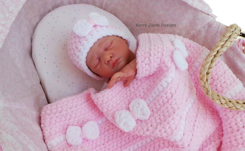 K&ouml;nnte beinhalten: Eine rosa geh&auml;kelte Babydecke mit wei&szlig;en geh&auml;kelten Blumen und Schleifen. Ein Baby schl&auml;ft auf der Decke und tr&auml;gt eine rosa und wei&szlig;e geh&auml;kelte M&uuml;tze mit einer wei&szlig;en Schleife.