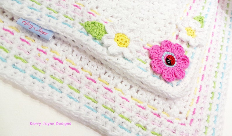 May include: White crochet baby blanket with a colorful striped border and flower appliques. The blanket features a pink flower with a ladybug button in the center. The label "Kerry Jayne Designs" is visible in the top left corner.