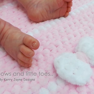 K&ouml;nnte beinhalten: Die F&uuml;&szlig;e eines Babys auf einer rosa und wei&szlig;en H&auml;keldecke mit wei&szlig;en Schleifen. Die Decke hat eine flauschige Textur.