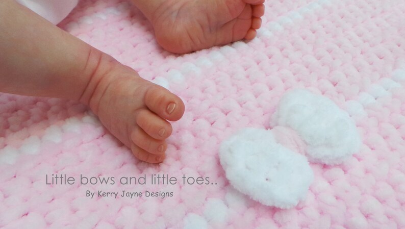 K&ouml;nnte beinhalten: Die F&uuml;&szlig;e eines Babys auf einer rosa und wei&szlig;en H&auml;keldecke mit wei&szlig;en Schleifen. Die Decke hat eine flauschige Textur.