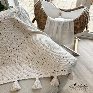 May include: A cream-colored, crocheted baby blanket with a diamond pattern and tassel trim. The blanket is draped over a chair and a baby is in a wicker bassinet in the background. The blanket is handmade and has a soft, textured appearance.