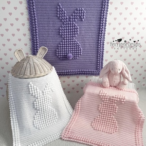 Puede incluir: Tres mantas de bebé de ganchillo con un diseño de conejo. Las mantas son de color morado, blanco y rosa. Las mantas están sobre una superficie blanca con un patrón de corazones rosas.