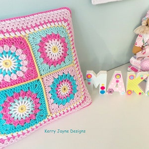 May include: A square crocheted pillow with a pink, white, yellow, and blue floral pattern. The pillow is sitting on a white surface with the words "Kerry Jayne Designs" printed below it.