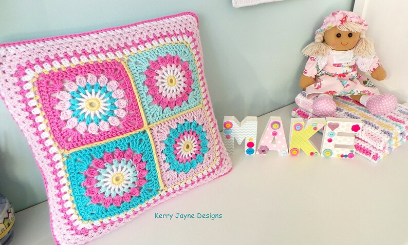 May include: A square crocheted pillow with a pink, white, yellow, and blue floral pattern. The pillow is sitting on a white surface with the words "Kerry Jayne Designs" printed below it.