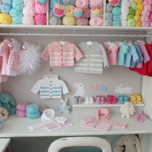 May include: A collection of crocheted baby sweaters and hats in various colors, including pink, blue, white, and gray. The sweaters have striped patterns and button closures. The hats have a hooded design. The image also includes a variety of yarn balls and crocheted owls.