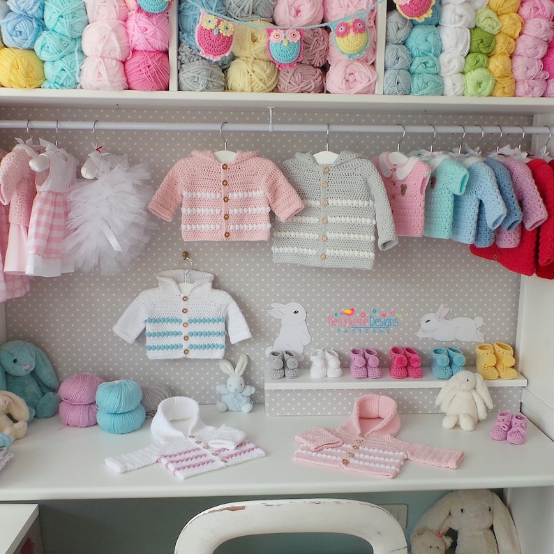 May include: A collection of crocheted baby sweaters and hats in various colors, including pink, blue, white, and gray. The sweaters have striped patterns and button closures. The hats have a hooded design. The image also includes a variety of yarn balls and crocheted owls.