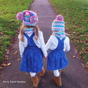 May include: Two children wearing matching blue denim overalls and white shirts walk down a path. They are both wearing colorful crocheted hats with pom-poms.