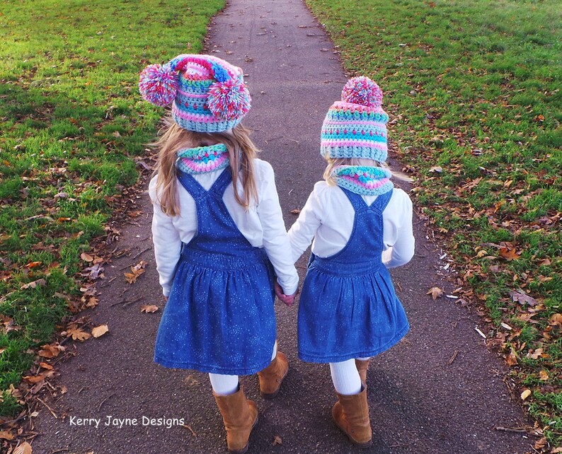 May include: Two children wearing matching blue denim overalls and white shirts walk down a path. They are both wearing colorful crocheted hats with pom-poms.