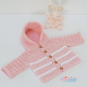 May include: A pink crocheted baby jacket with a hood and white stripes. The jacket has wooden buttons and is laid out on a white surface.