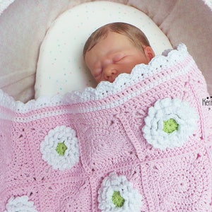 May include: A pink crocheted baby blanket with white flowers and a green center. The blanket has a white crocheted border. The blanket is in a white bassinet with a pink and white polka dot lining.