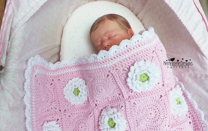 May include: A pink crocheted baby blanket with white flowers and a green center. The blanket has a white crocheted border. The blanket is in a white bassinet with a pink and white polka dot lining.