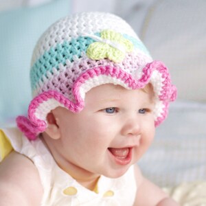 Puede incluir: Un beb&eacute; con un gorro de ganchillo rosa, blanco y azul con un adorno de flor verde. El gorro tiene un borde rizado.