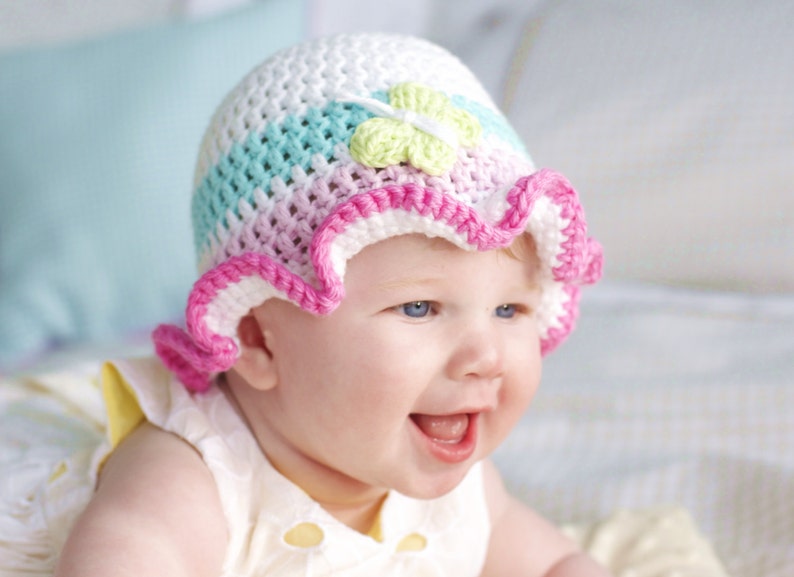 Puede incluir: Un beb&eacute; con un gorro de ganchillo rosa, blanco y azul con un adorno de flor verde. El gorro tiene un borde rizado.