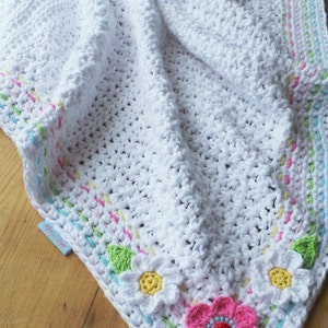 May include: A white crocheted baby blanket with a colorful border and flower appliques. The blanket is made with a soft, textured yarn and features a ladybug in the center of one of the flowers. The blanket is labeled "The 'Little Ladybird Blanket' By Kerry Jayne Designs."