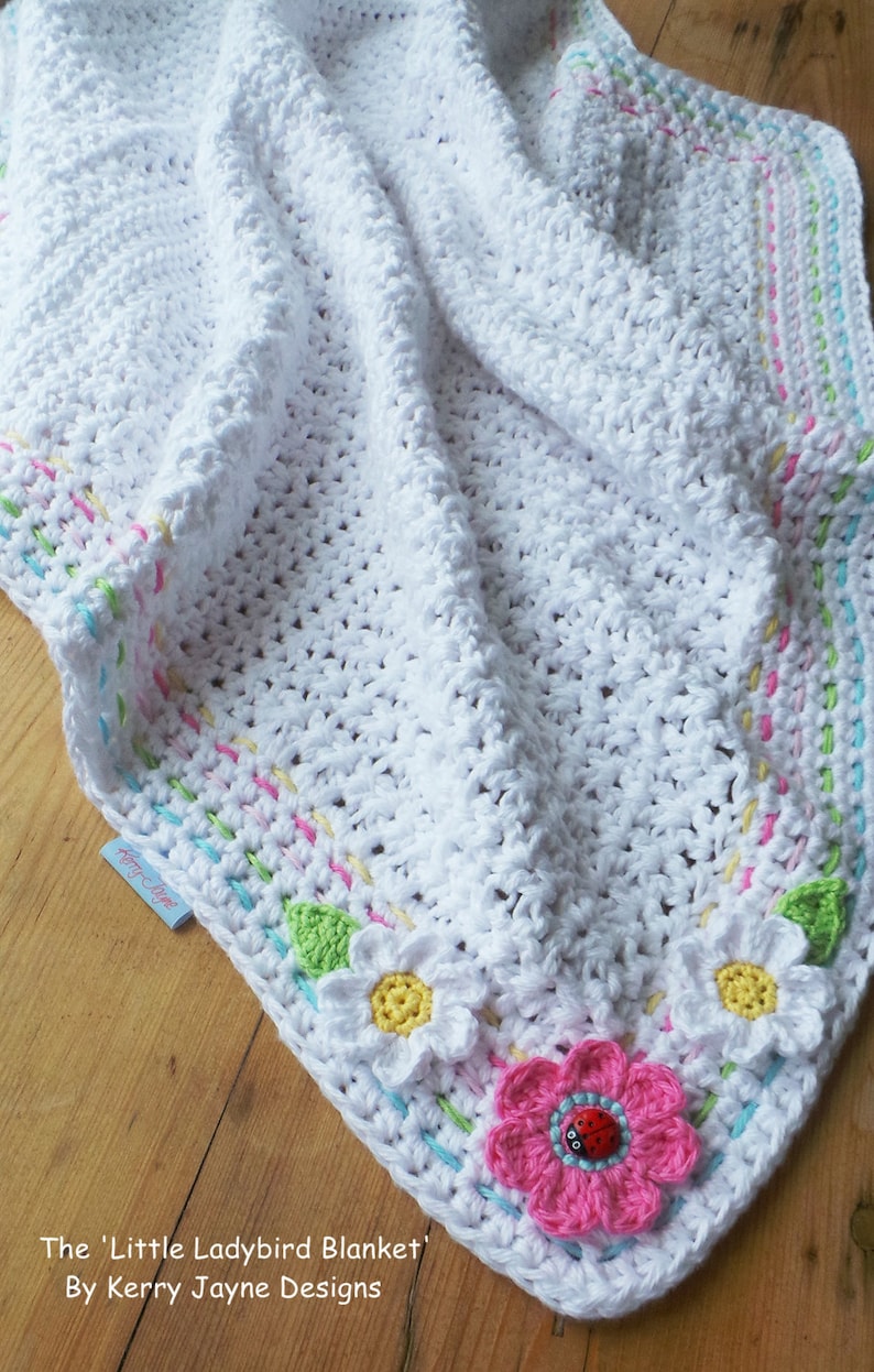 May include: A white crocheted baby blanket with a colorful border and flower appliques. The blanket is made with a soft, textured yarn and features a ladybug in the center of one of the flowers. The blanket is labeled "The 'Little Ladybird Blanket' By Kerry Jayne Designs."