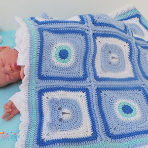 K&ouml;nnte beinhalten: Eine blaue und wei&szlig;e geh&auml;kelte Babydecke mit einem B&auml;renmuster. Die Decke ist aus verschiedenen Blaut&ouml;nen und wei&szlig;em Garn gefertigt und hat einen Rand aus wei&szlig;em und hellblauem Garn.