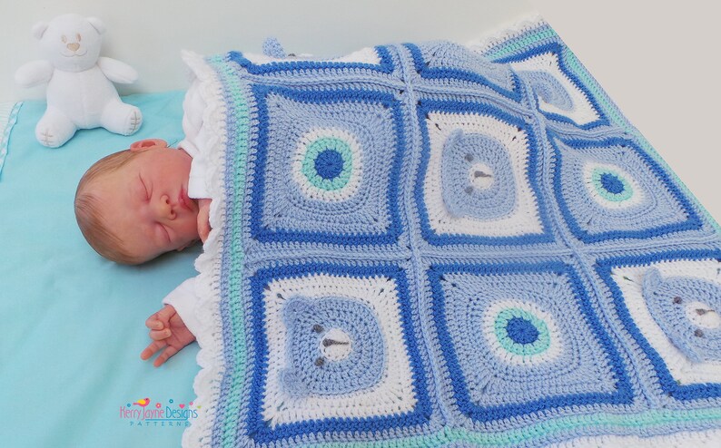 K&ouml;nnte beinhalten: Eine blaue und wei&szlig;e geh&auml;kelte Babydecke mit einem B&auml;renmuster. Die Decke ist aus verschiedenen Blaut&ouml;nen und wei&szlig;em Garn gefertigt und hat einen Rand aus wei&szlig;em und hellblauem Garn.