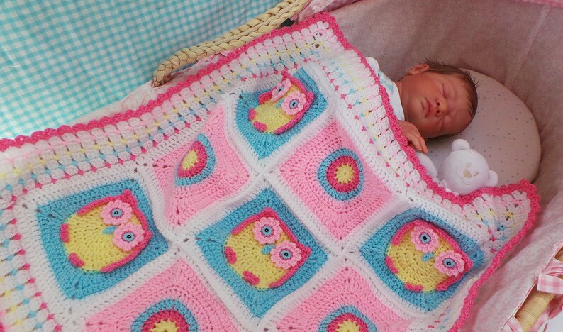 May include: A pink, blue, and white crocheted baby blanket with a scalloped edge and owl designs. The blanket is in a wicker basket with a baby doll sleeping on top.