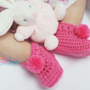 Puede incluir: Botas de beb&eacute; de ganchillo rosa con pompones en la parte superior. Las botas est&aacute;n sobre una superficie blanca.