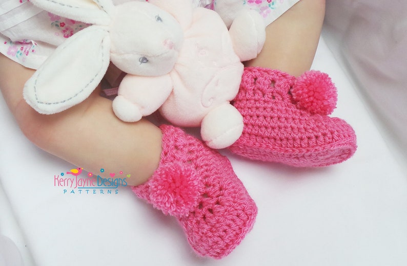 Puede incluir: Botas de beb&eacute; de ganchillo rosa con pompones en la parte superior. Las botas est&aacute;n sobre una superficie blanca.
