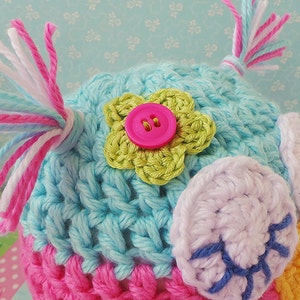 May include: A crocheted owl hat with pink, blue, and white yarn. The hat has a green flower with a pink button in the center and white crocheted eyes.