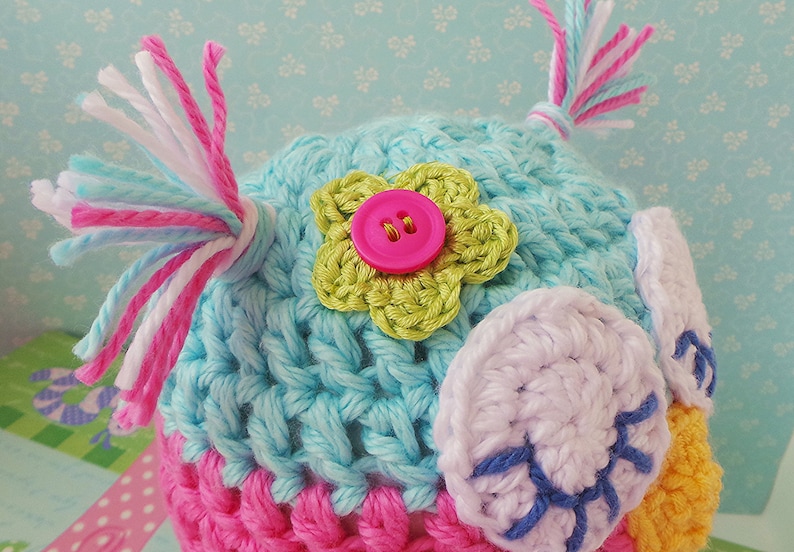 May include: A crocheted owl hat with pink, blue, and white yarn. The hat has a green flower with a pink button in the center and white crocheted eyes.