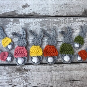May include: Crocheted bunny butt ornaments in various colors. Each bunny has gray ears, a white fluffy tail, and a colorful bottom in shades of pink, yellow, orange, and green. The ornaments are arranged on a weathered wooden surface.