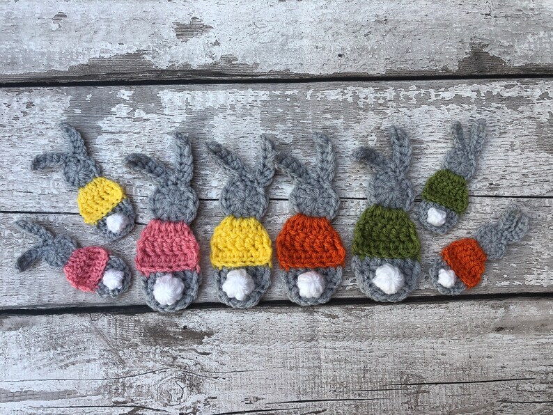 May include: Crocheted bunny butt ornaments in various colors. Each bunny has gray ears, a white fluffy tail, and a colorful bottom in shades of pink, yellow, orange, and green. The ornaments are arranged on a weathered wooden surface.