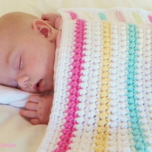 Peut inclure: Un b&eacute;b&eacute; dort paisiblement sous une couverture de b&eacute;b&eacute; en crochet blanc avec des rayures roses, jaunes et vertes.