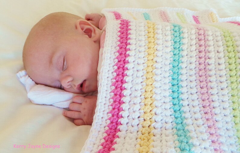 Peut inclure: Un b&eacute;b&eacute; dort paisiblement sous une couverture de b&eacute;b&eacute; en crochet blanc avec des rayures roses, jaunes et vertes.