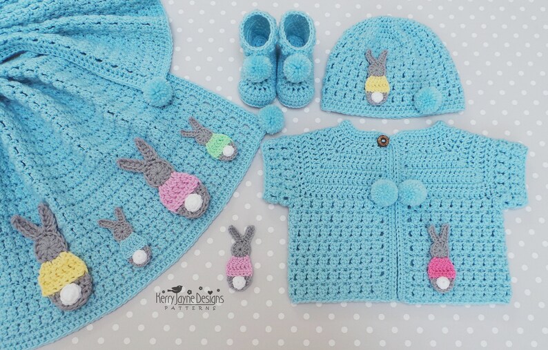 May include: A light blue crochet baby set featuring a blanket, booties, hat, and cardigan. Each item is adorned with gray, pink, yellow, and white bunny appliqu&eacute;s and pom-poms. The set is handmade and suitable for a baby.