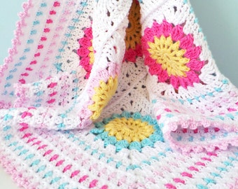 May include: A crocheted baby blanket with a white background and colorful flowers in shades of pink, yellow, and blue. The blanket has a scalloped edge.