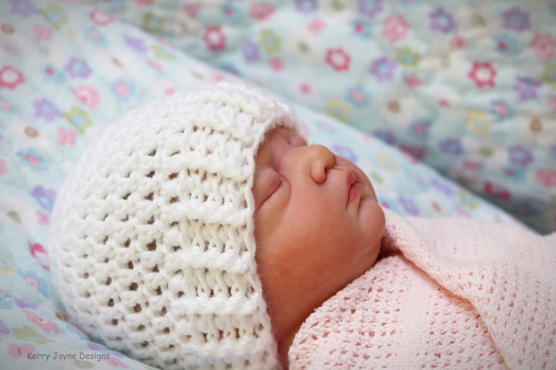 May include: A white crocheted hat with a ribbed texture sits on the head of a sleeping baby. The baby is wearing a pink crocheted blanket.