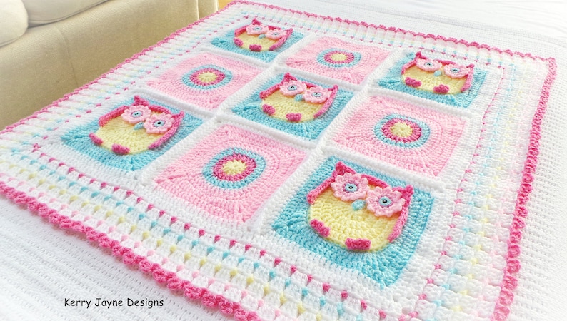 May include: A crocheted baby blanket with a pink, yellow, and blue color scheme. The blanket features a repeating pattern of owls and circles. The blanket is edged with a pink crocheted border. The text "Kerry Jayne Designs" is visible in the lower left corner of the image.