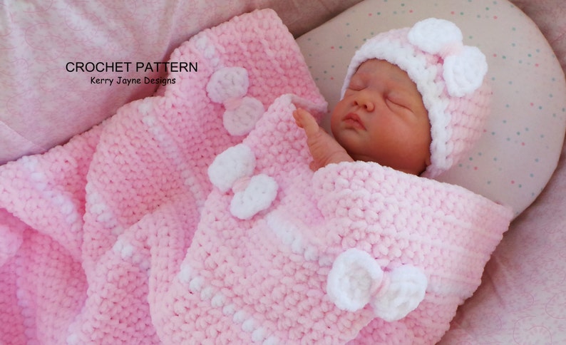 K&ouml;nnte beinhalten: Eine rosa geh&auml;kelte Babydecke mit wei&szlig;en geh&auml;kelten Blumen und einer passenden rosa-wei&szlig;en geh&auml;kelten M&uuml;tze.
