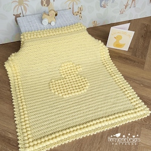 Puede incluir: Una manta de bebé de crochet amarilla con un diseño de pato de goma texturizado en el centro. La manta tiene un borde de pequeñas bolas amarillas. El texto "Bienvenido pequeño" es visible en una tarjeta en el fondo.