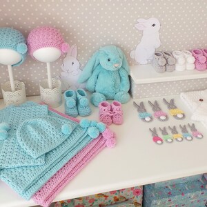 May include: A display of handmade baby items, including crochet hats, booties, blankets, and bunny-shaped appliques. The color palette includes blue, pink, white, and yellow. A plush bunny and decorative elements complete the scene.