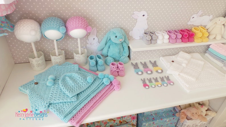May include: A display of handmade baby items, including crochet hats, booties, blankets, and bunny-shaped appliques. The color palette includes blue, pink, white, and yellow. A plush bunny and decorative elements complete the scene.