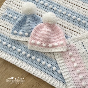 Puede incluir: Dos gorros de bebé de ganchillo, uno azul y otro rosa, con pompones blancos y ribete de ganchillo blanco. Los gorros están encima de una manta de bebé de ganchillo con rayas blancas y azules.