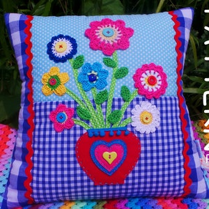 May include: A blue and white gingham cushion with a red scalloped border. The cushion features a crocheted flower arrangement in a red heart-shaped vase. The flowers are pink, yellow, white, and blue. The text "PATTERN PDF" is printed on the right side of the cushion.