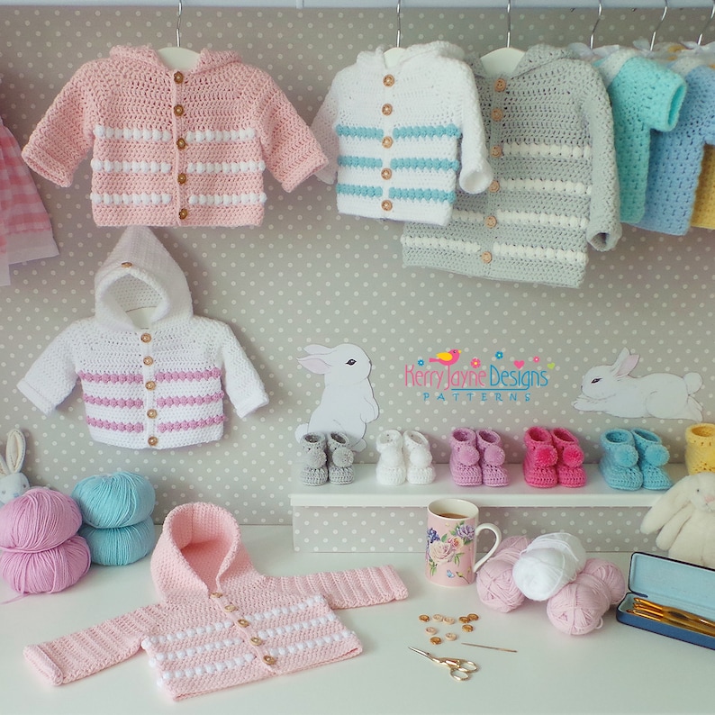 K&ouml;nnte beinhalten: Eine Sammlung von geh&auml;kelten Babyjacken in Rosa, Blau und Grau mit wei&szlig;en Streifen und Kn&ouml;pfen. Die Jacken h&auml;ngen an Kleiderb&uuml;geln und auf einem Tisch darunter befinden sich Wollkn&auml;uel, H&auml;kelnadeln und andere Bastelutensilien.